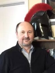A professor in a black sweater sitting in front of a Roman era helmet.